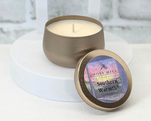 8 oz Soy Candle in the scent southern warmth. Hand-poured into a gold tin. Sitting on top of a white table.