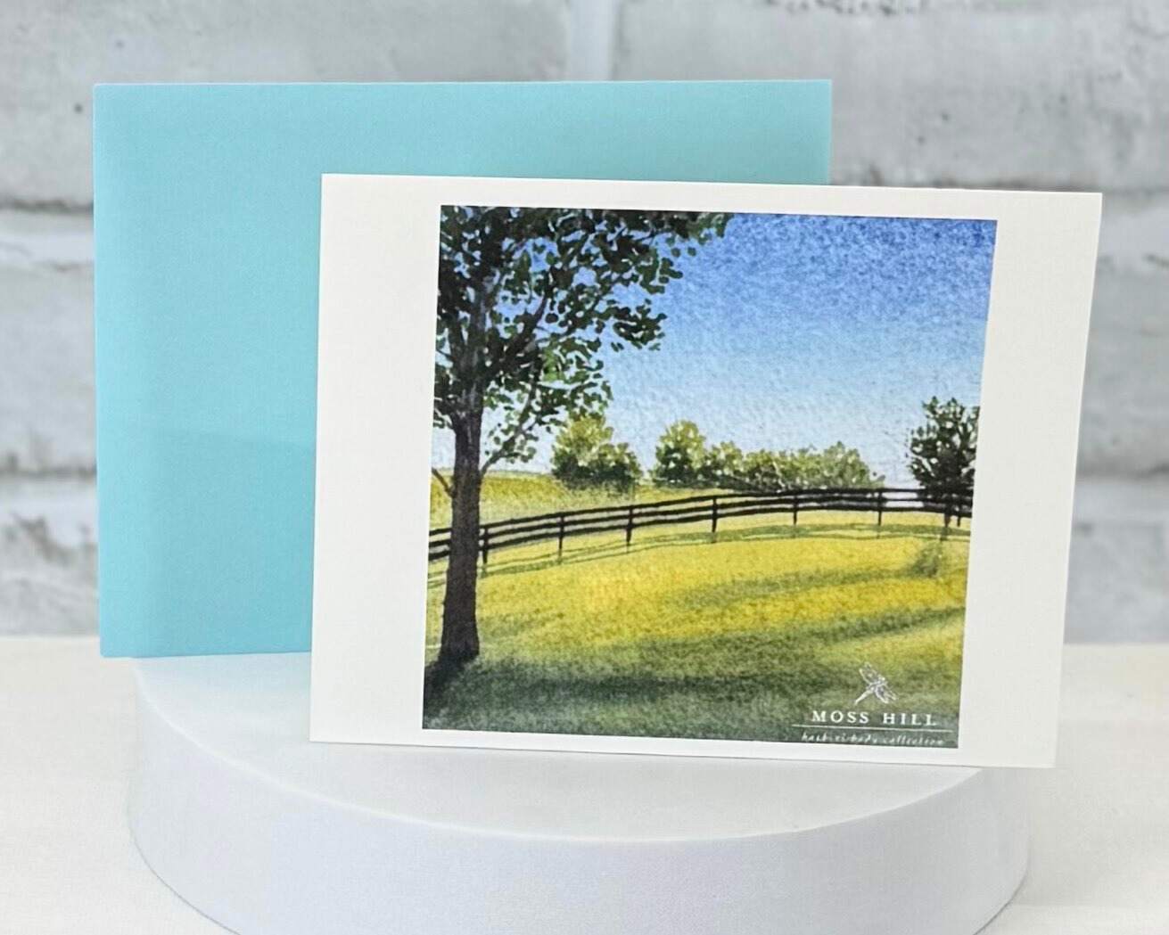 Card with a scenic watercolor painting of a field and trees on a white stand with a light blue card behind it. This represents the Moss Hill Bluegrass scent.
