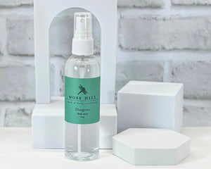 Bluegrass body spray, handmade by Moss Hill. In a clear container sitting on a white tabletop.