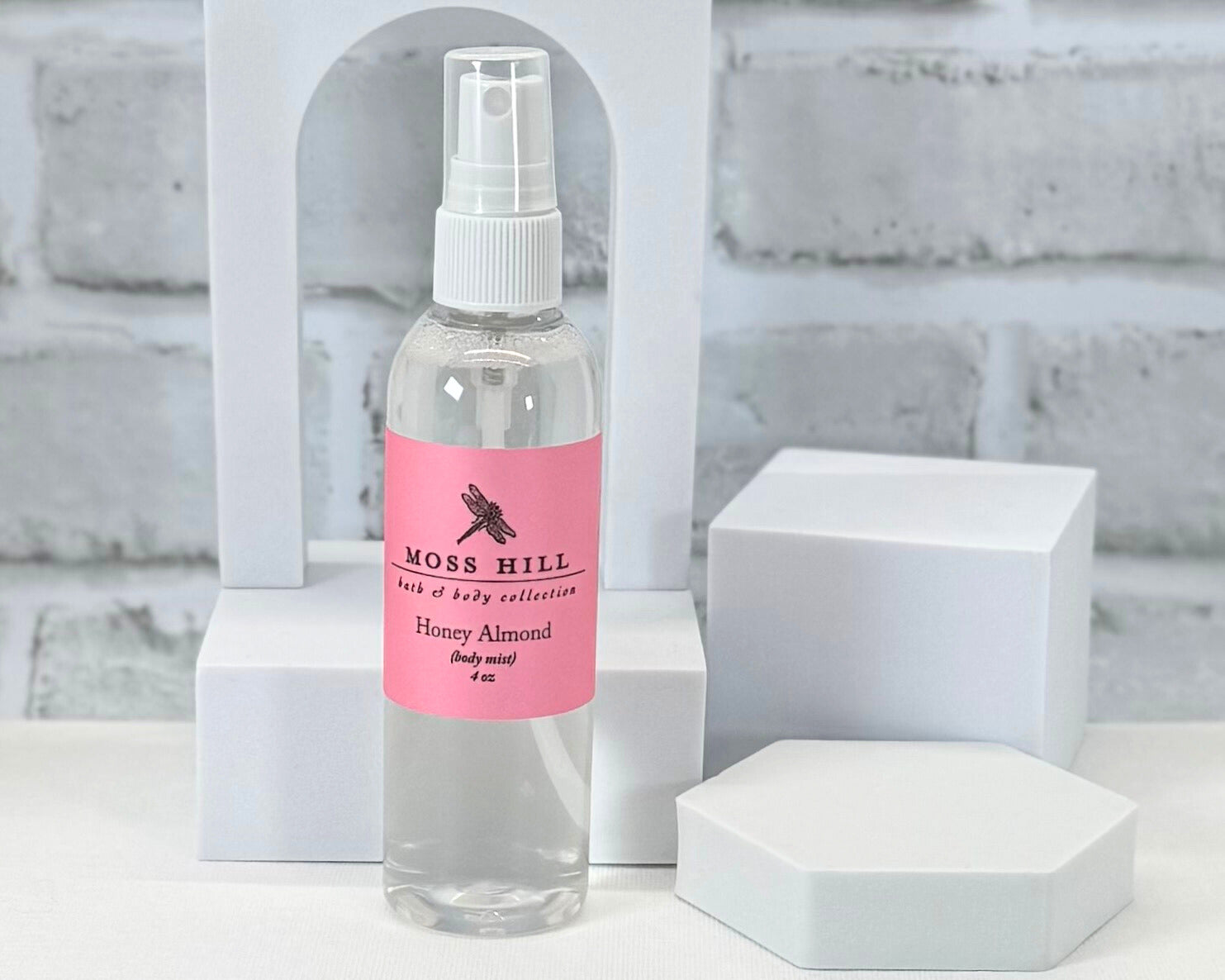 Honey Almond Body Mist. In a clear container with a pink label with the Moss Hill Bath & Body Collection Logo.