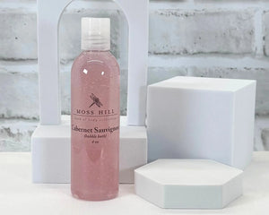 A pink bottle of handmade bubble bath in the scent Cabernet Sauvignon. Place on a white surface with a white background.