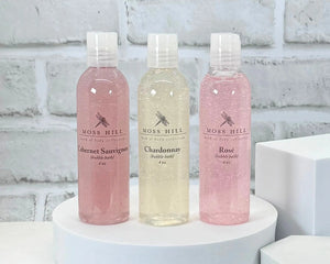 Three bottles of Moss Hill handmade bubble bath on a white platform with a light gray brick wall background.