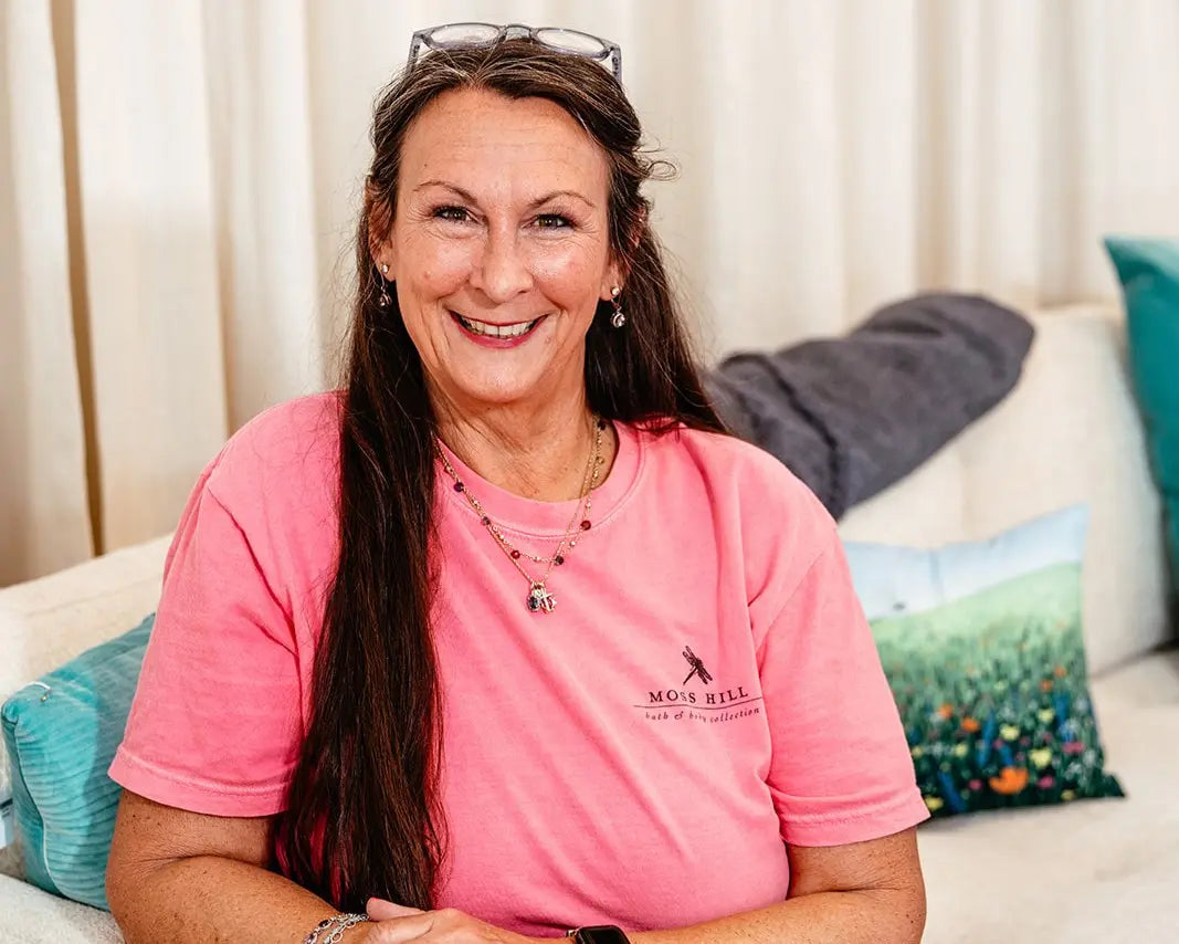 Woman wearing a pink Moss Hill Flutter shirt with a logo, sitting on a couch with colorful pillows.