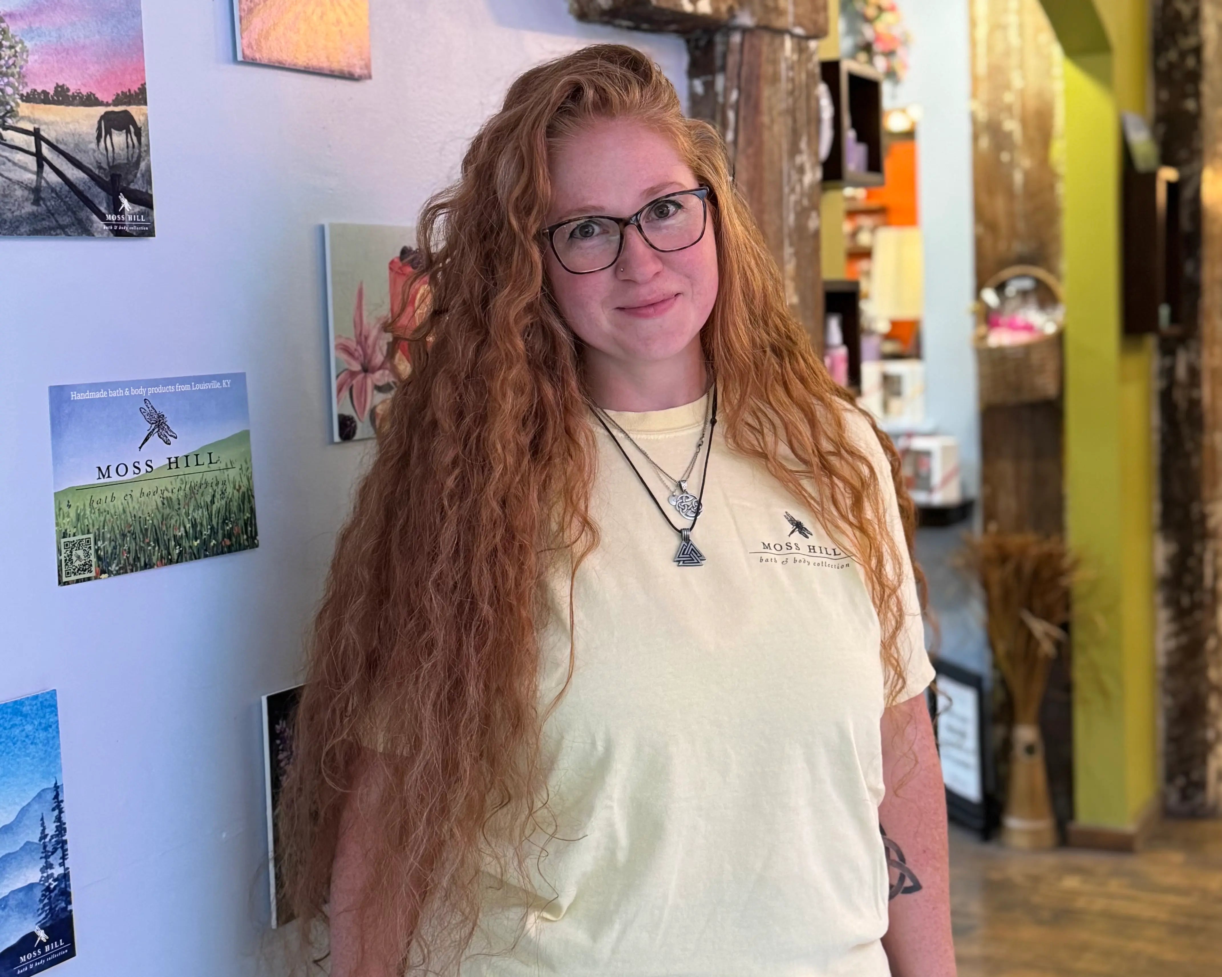 Person with long red hair wearing glasses and a yellow Moss Hill Flutter shirt in an indoor setting.