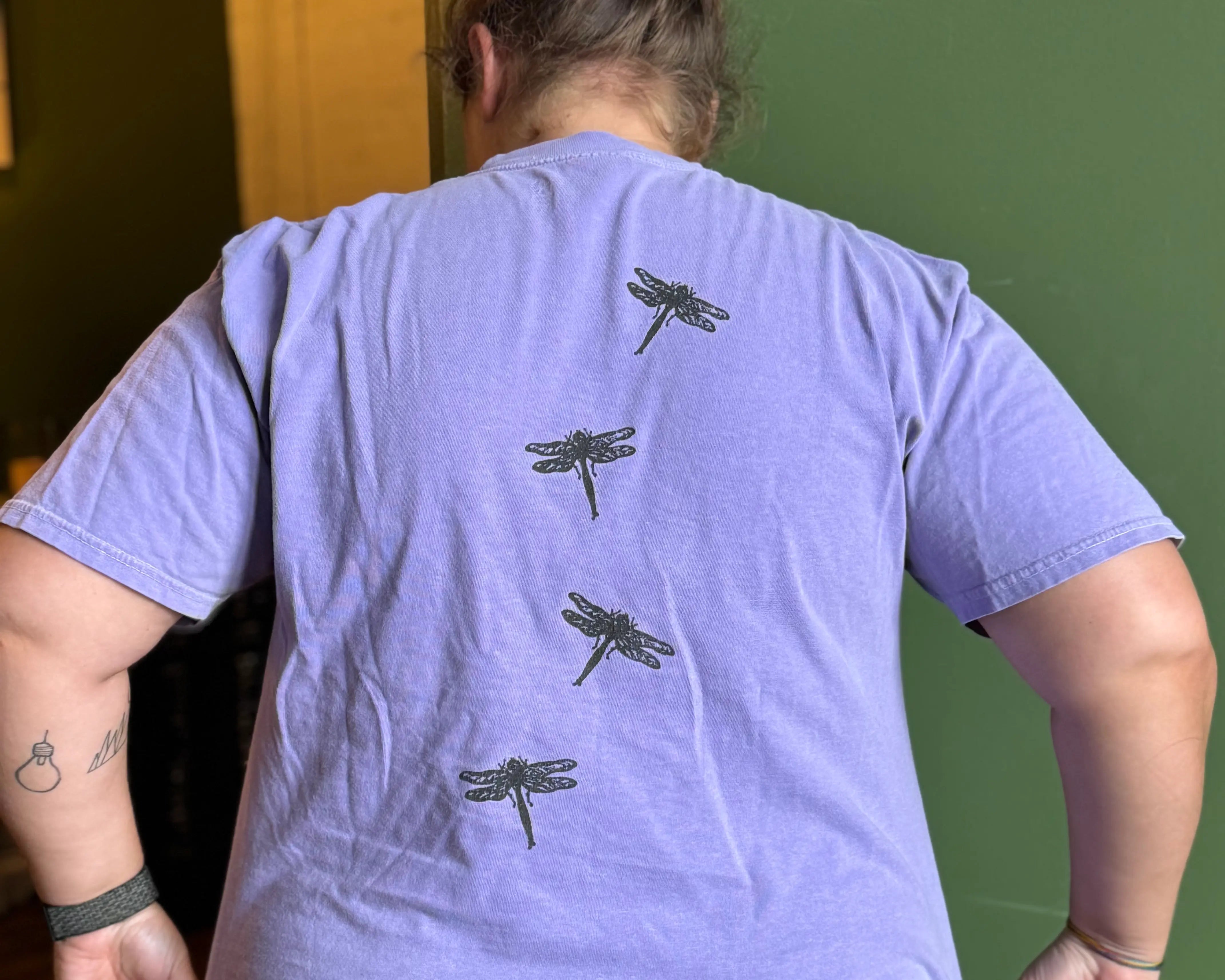 Person wearing a purple t-shirt with Moss Hill dragonfly designs on the back, standing against a green wall.