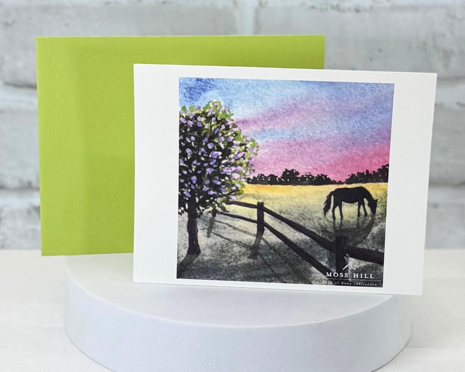 A watercolor greeting card with a horse and field illustration on a white stand with a green envelope. Representing the Kentucky Girl scented collection at Moss Hill Bath and Body.