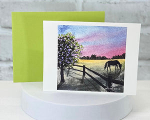 A watercolor greeting card with a horse and field illustration on a white stand with a green envelope. Representing the Kentucky Girl scented collection at Moss Hill Bath and Body.
