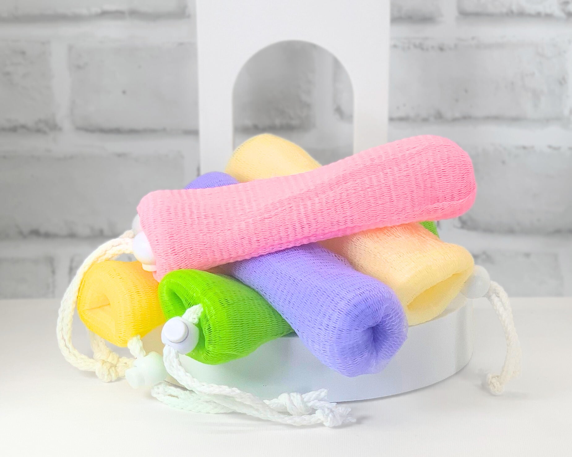 A collection of colorful soap loofas made of mesh, tied with drawstrings, presented in a variety of colors including tan, green, purple, pink, and yellow.