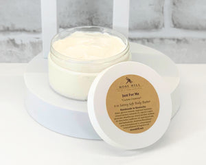 A jar of body butter with the lid off, sitting on a white surface. The label on the lid reads 'Just For Me' and indicates the product is a 6 oz Satiny Soft Body Butter.