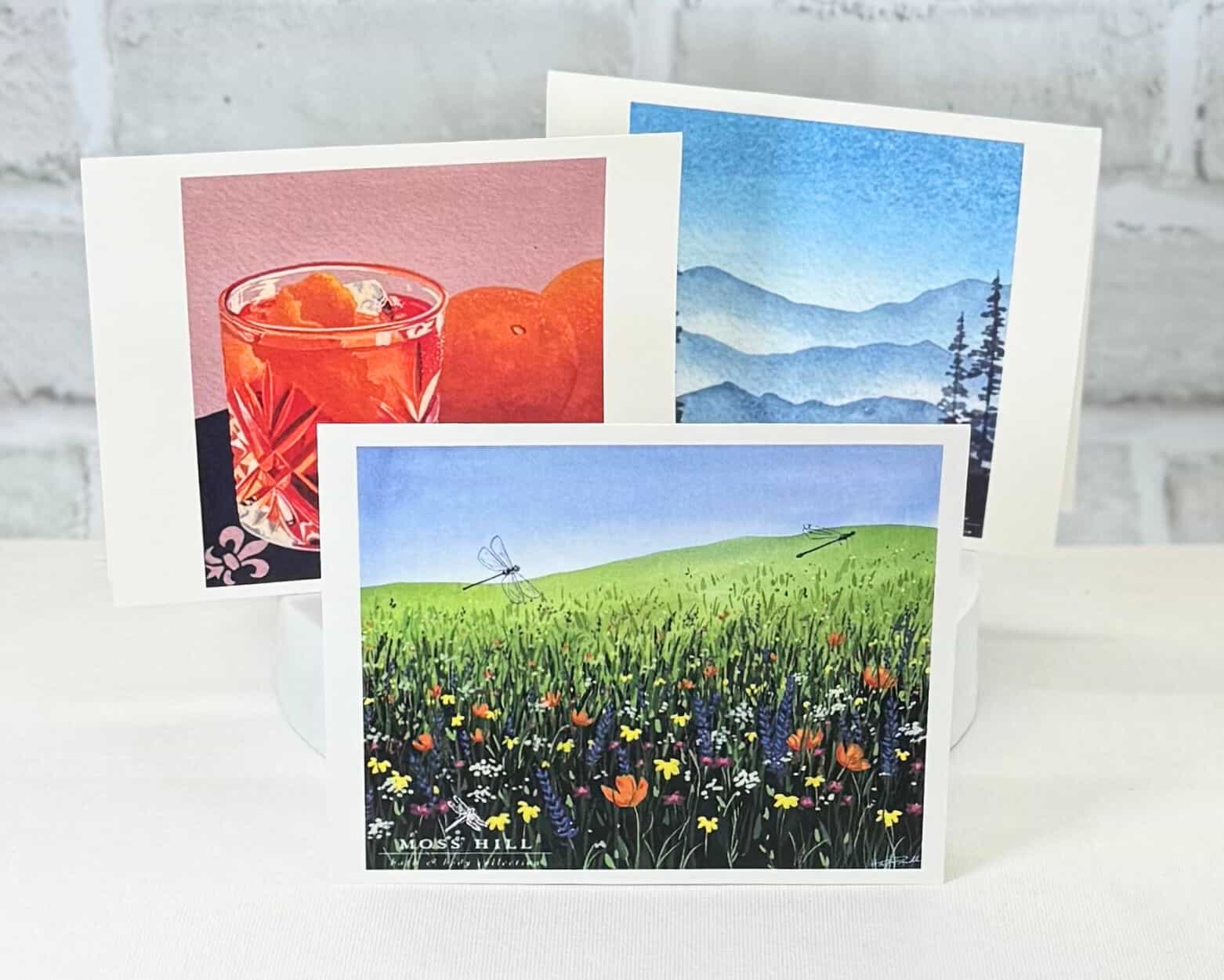 Three greeting cards with different watercolor illustrations on a white surface. Representing Moss Hill's signature scents.
