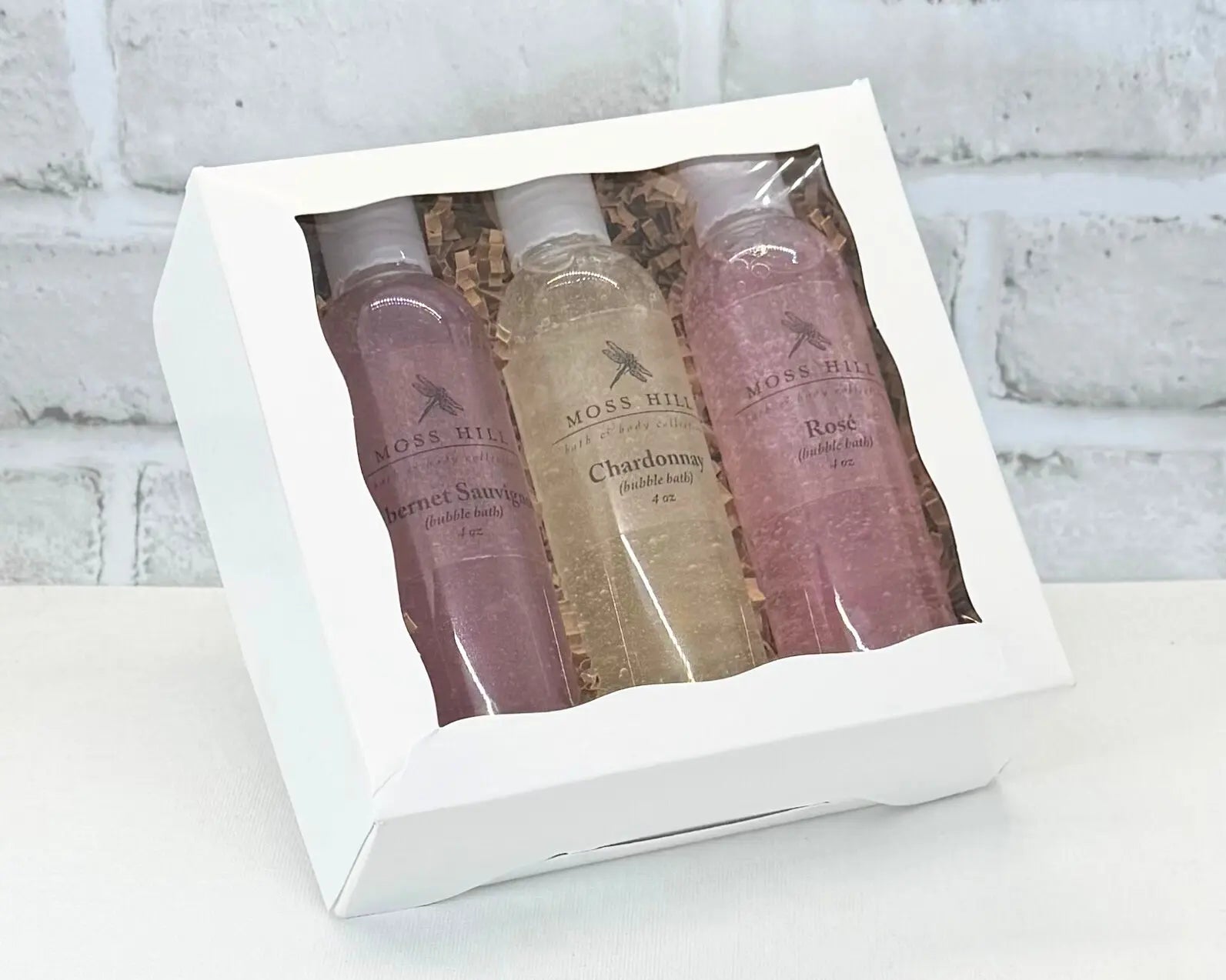 Three bottle of wine scented handmade bubble baths. Packed in a white box, sitting on a white surface, with a white brick background.
