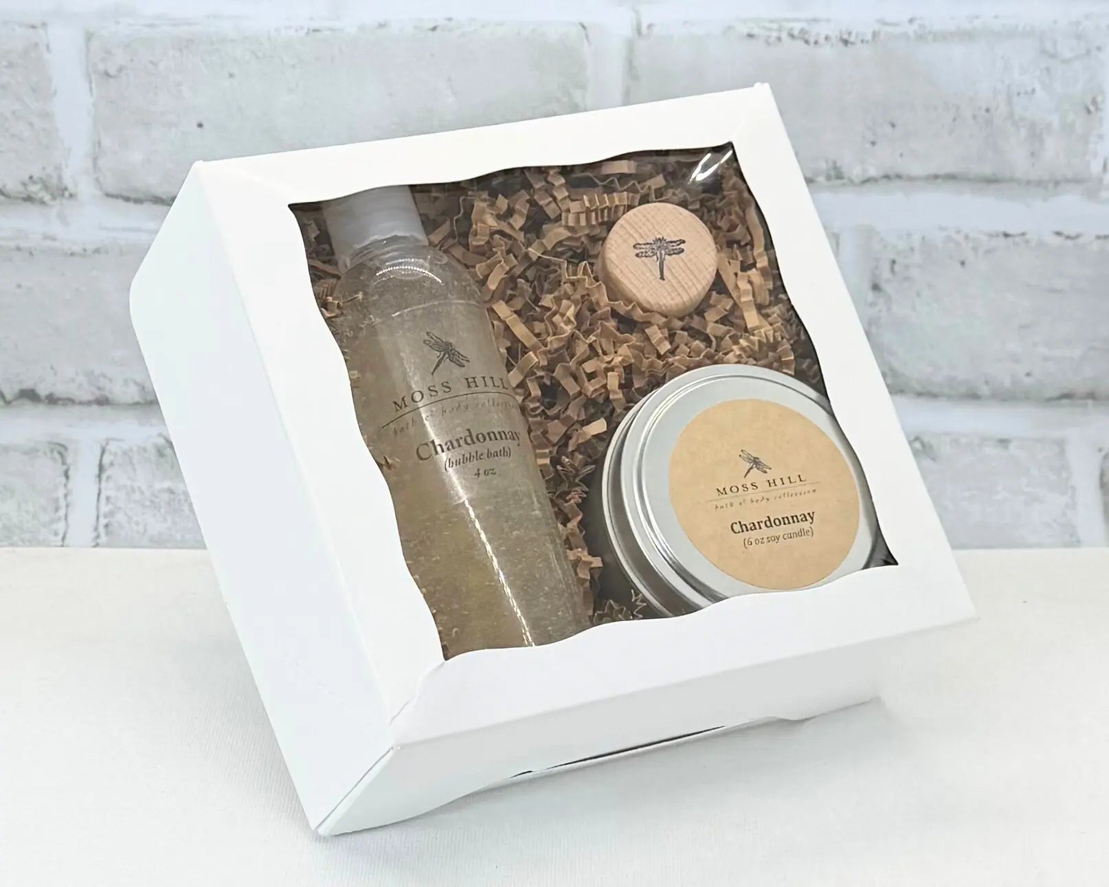 A Moss Hill body care gift set with a handmade bubble bath and soy candle in the scent Chardonnay. Wrapped in a white box placed on a surface with white brick background.
