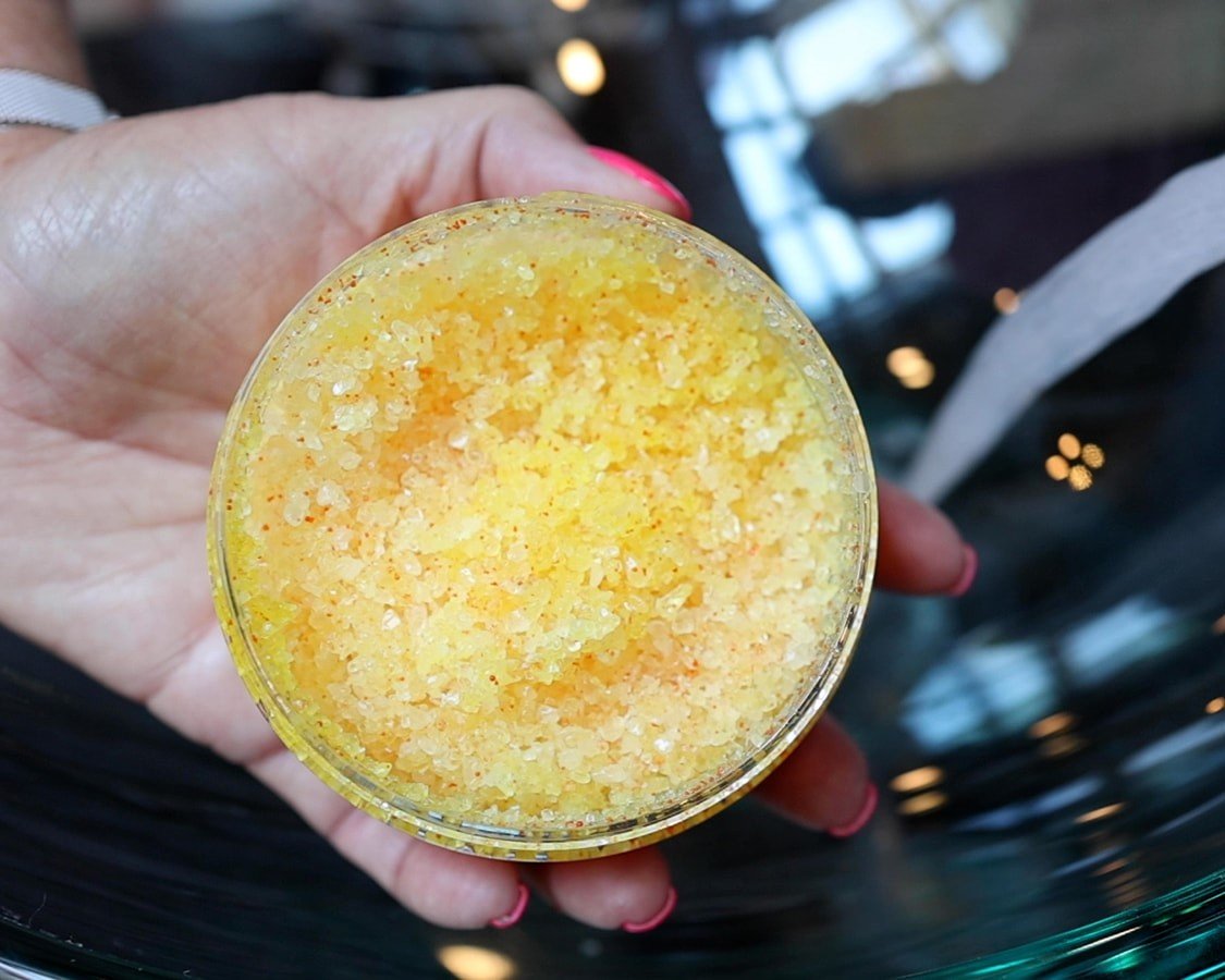 A clear container of yellow body scrub being held to show the body care product