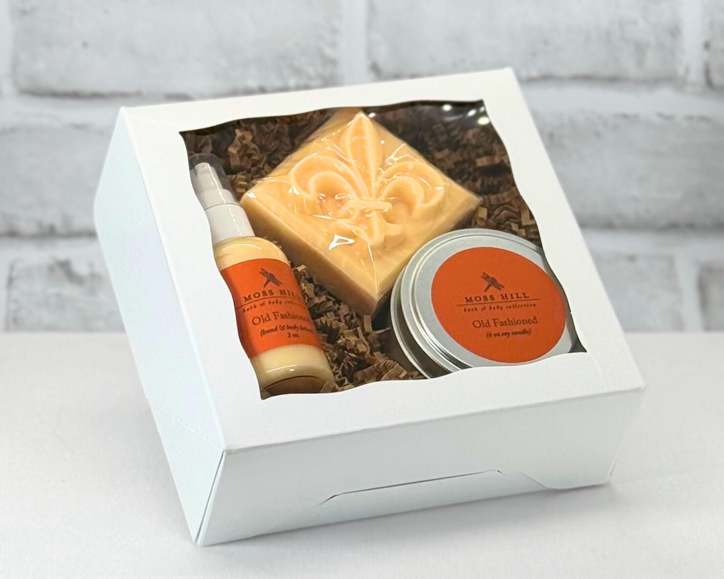3 piece body care gift box set with bar soap, hand a body lotion, and a 6 oz soy candle in the scent Old Fashioned. Wrapped in white box wit brown filler paper.