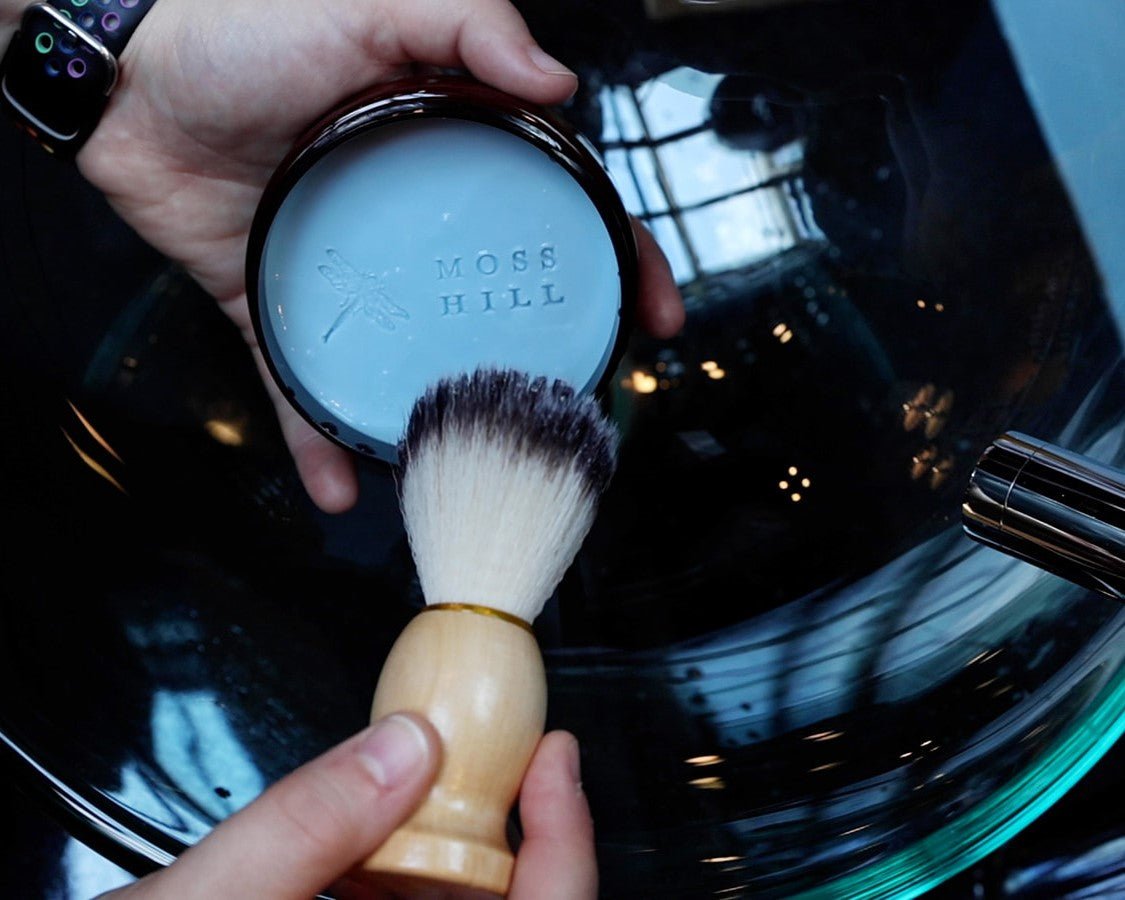 A shave brush being held next to handmade shave cream in a container with Moss Hill embossed details over a clear bowl sink.