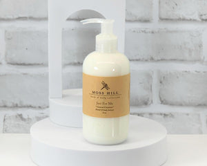 A white pump bottle of Moss Hill hand and body lotion placed on a white surface with a brick wall in the background. To symbolize creating your own custom hand & body lotion.