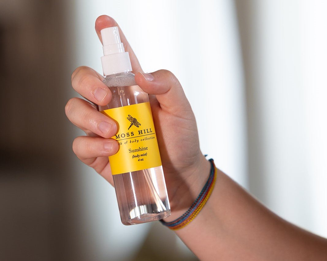 A clear bottle and white mister with a yellow label being held in one hand to represent spraying body mist or body spray