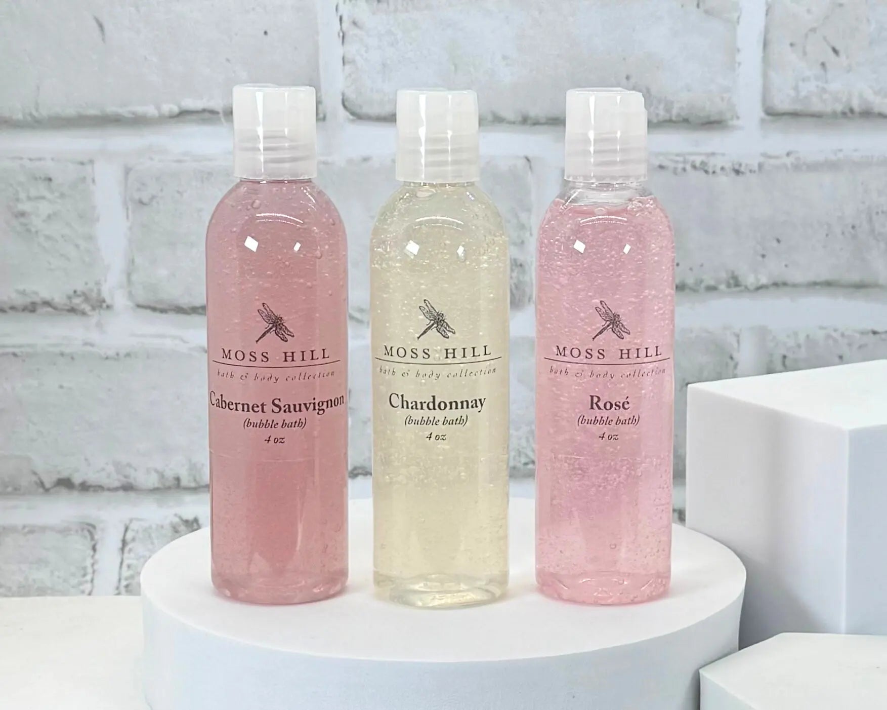 Three bottles of Moss Hill handmade bubble bath on a white platform with a light gray brick wall background.
