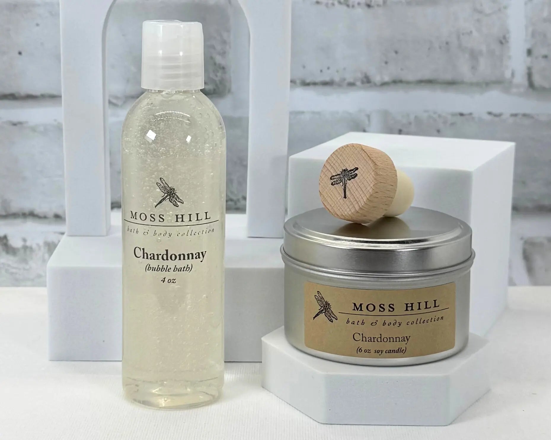 Moss Hill Chardonnay handmade bubble bath bottle and soy candle on a white surface with a gray brick wall background.