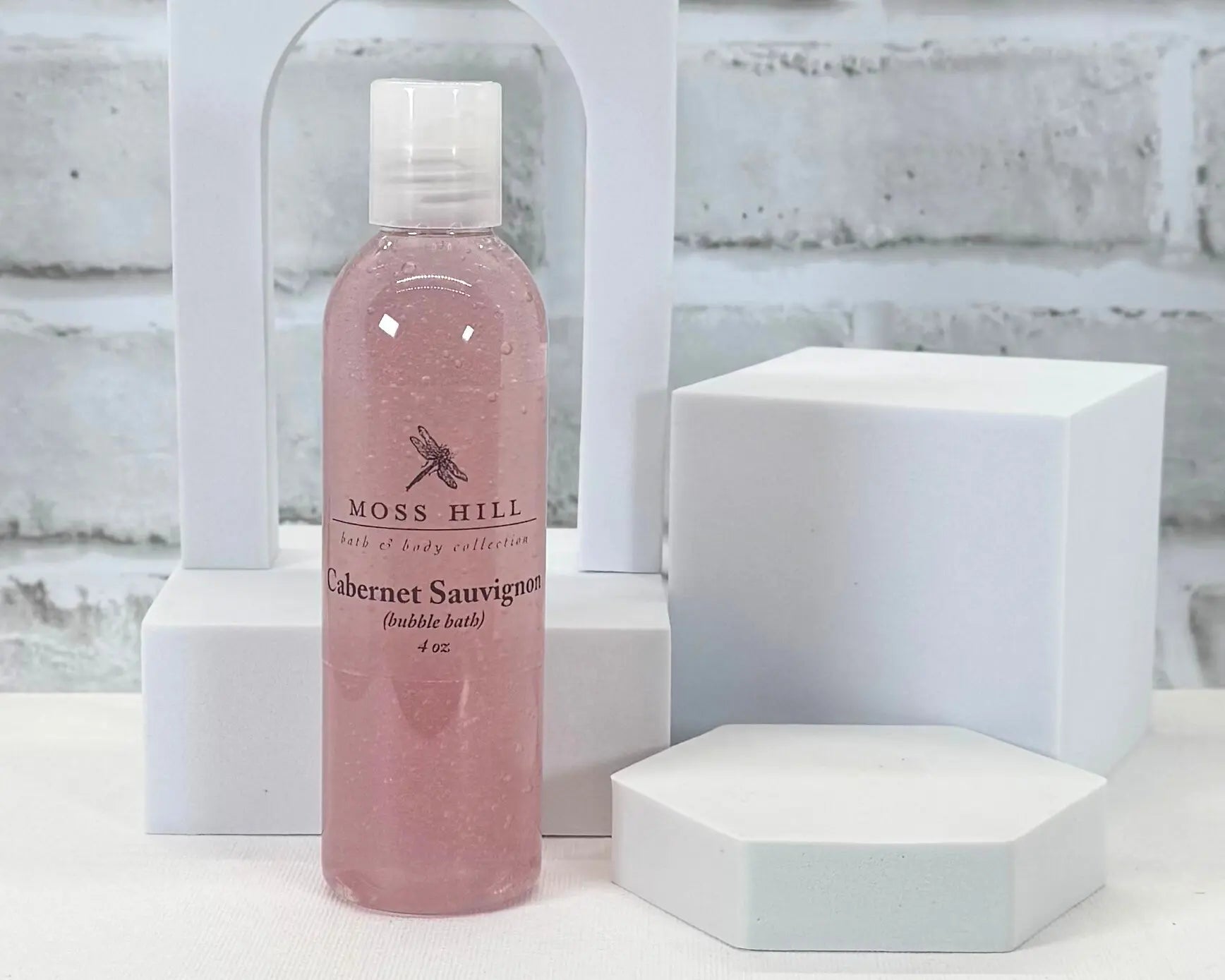 A pink bottle of handmade bubble bath in the scent Cabernet Sauvignon. Place on a white surface with a white background.