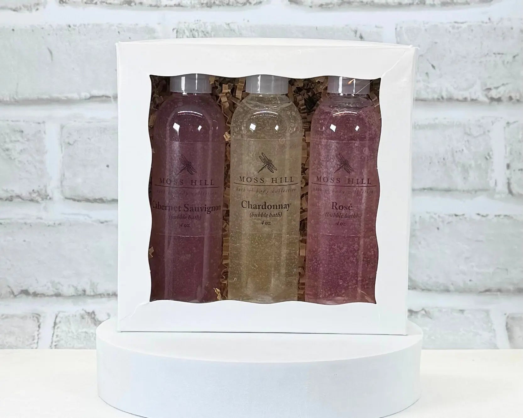 Moss Hill handmade bubble bath wine flight. 3 bottle of bubble batch packed in a white box, placed on a white surface, with a white brick background.