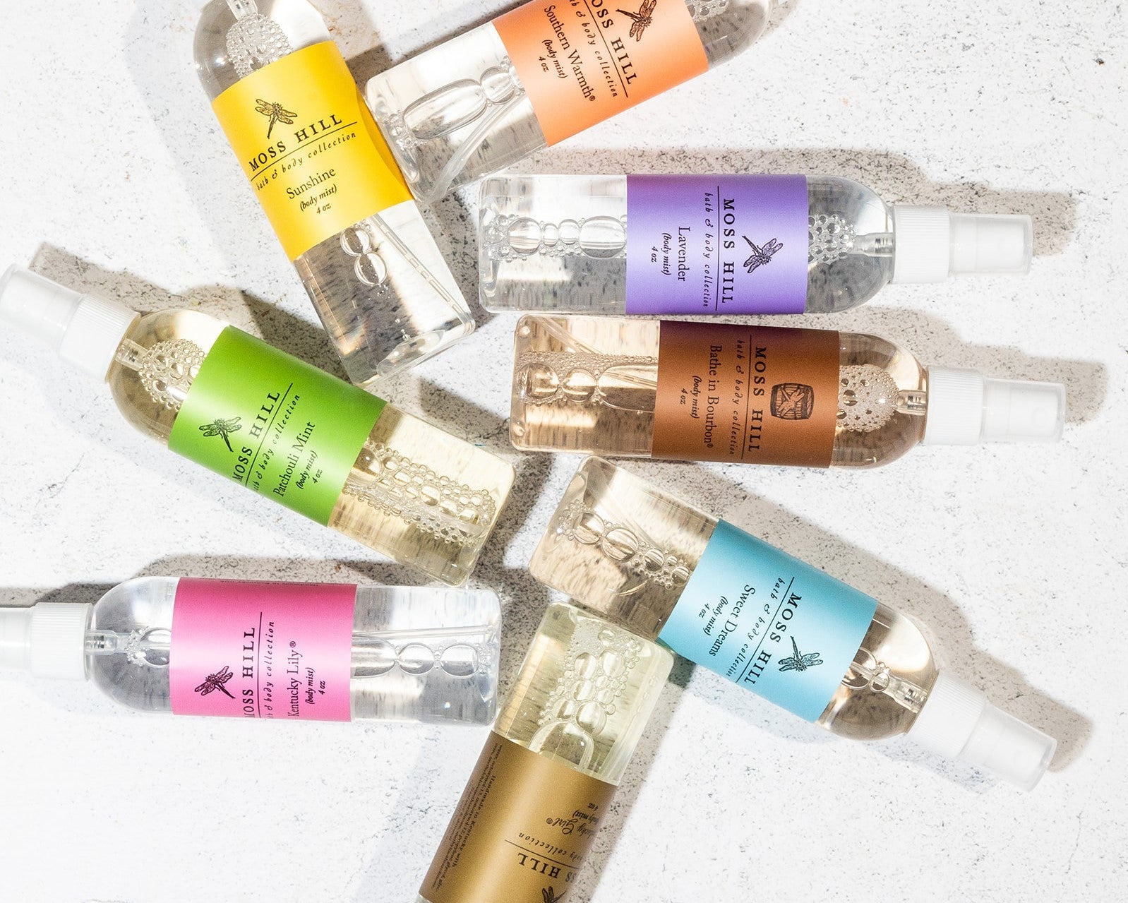 Several clear bottles with white sprayers of body mist laying on a white countertop in several different fragrances.