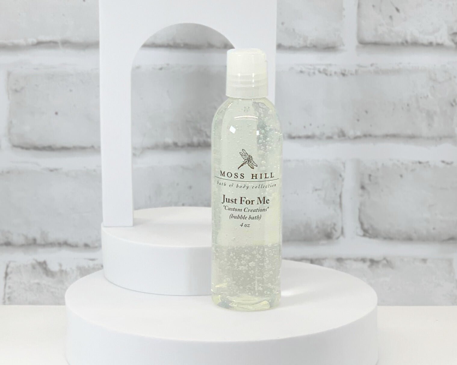 A clear plastic bottle of Moss Hill bubble bath on a white circular pedestal, with a white brick background. Labeled Moss Hill Bath & Body Collection, Just For Me, Custom Creations.