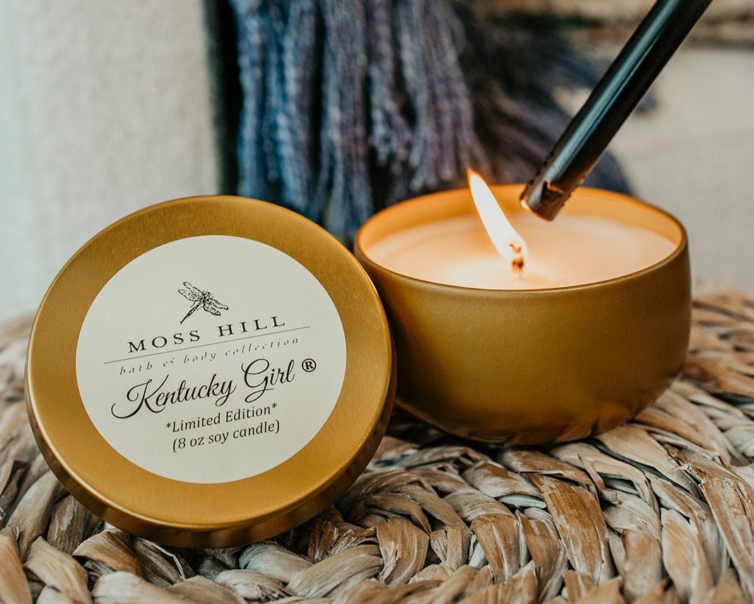 A soy candle in a gold tin with the white wick being lit by a long black lighter.