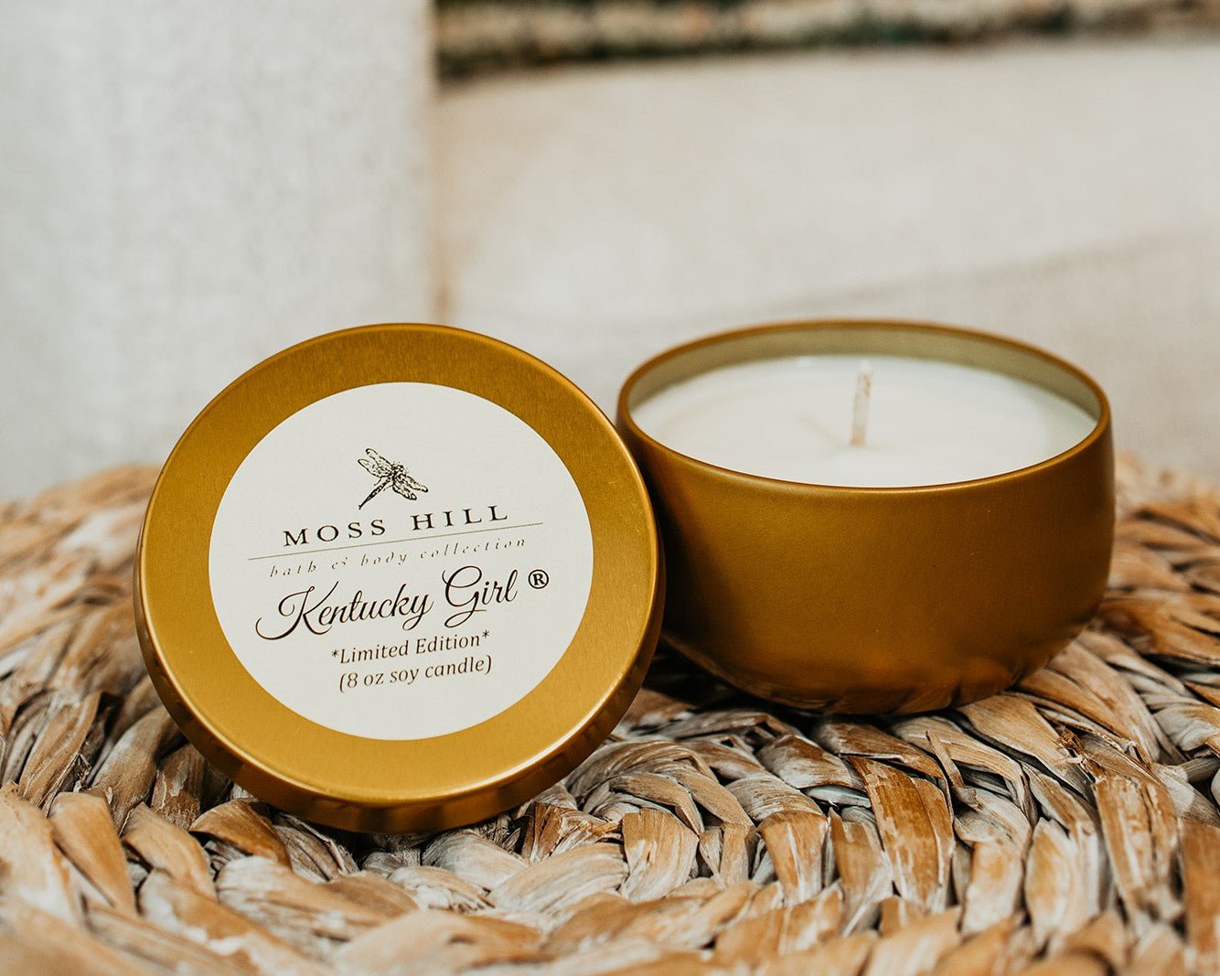 A gold lid to the side of the gold tin of hand poured soy candle with a white sick sitting on a woven basket.