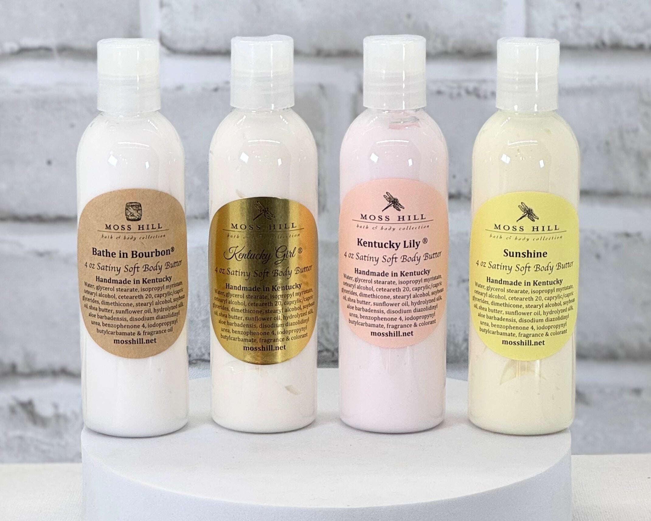 Four bottles of handmade body butter in a row on a white surface, with labels showing different scents such as 'Bathe in Bourbon', 'Kentucky Lily', and 'Sunshine'.