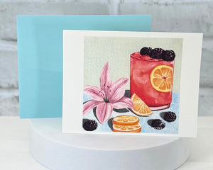 Greeting card with a drink illustration featuring a pink flower, blackberries, and oranges on a light blue background. Representing the Kentucky Lilly scented handmade bath and body products collection at Moss Hill