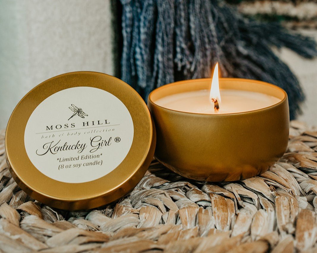 A lid of tin propped up against a lit soy candle in a gold tin sitting on a textured table setting
