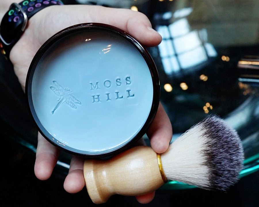 Hamdmade shave cream in a clear brown container being held in one hand with a shave brush
