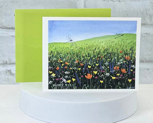 Greeting card with a field of flowers and dragonfly design on a white stand. Showcasing the scent Meadow. Which has handmade bath and body products for purchase.