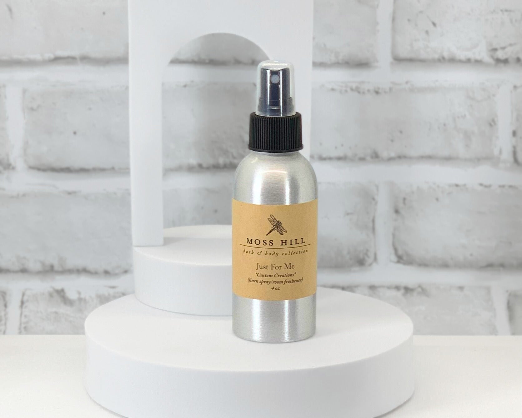 A silver linen spray bottle with a black pump standing on a white round surface with a white brick wall in the background. Labeled "Just for Me Custom Creations".