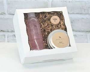 A Moss Hill body care gift set with handmade bubble bath, soy candle, and wine cork in the scent Rose. Packaged in a white box on a white brick background.