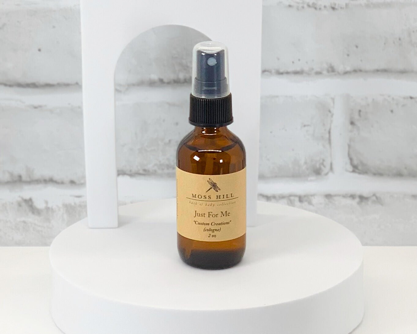 A brown spray bottle of Cologne labeled ' Moss Hill Custom Creations ' on a white surface with a brick wall in the background.