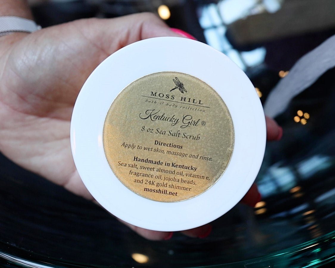 Hand holding body scrub with a white lid and gold sticker that read 8 oz os Sea Salt Scrub handmade in Kentucky