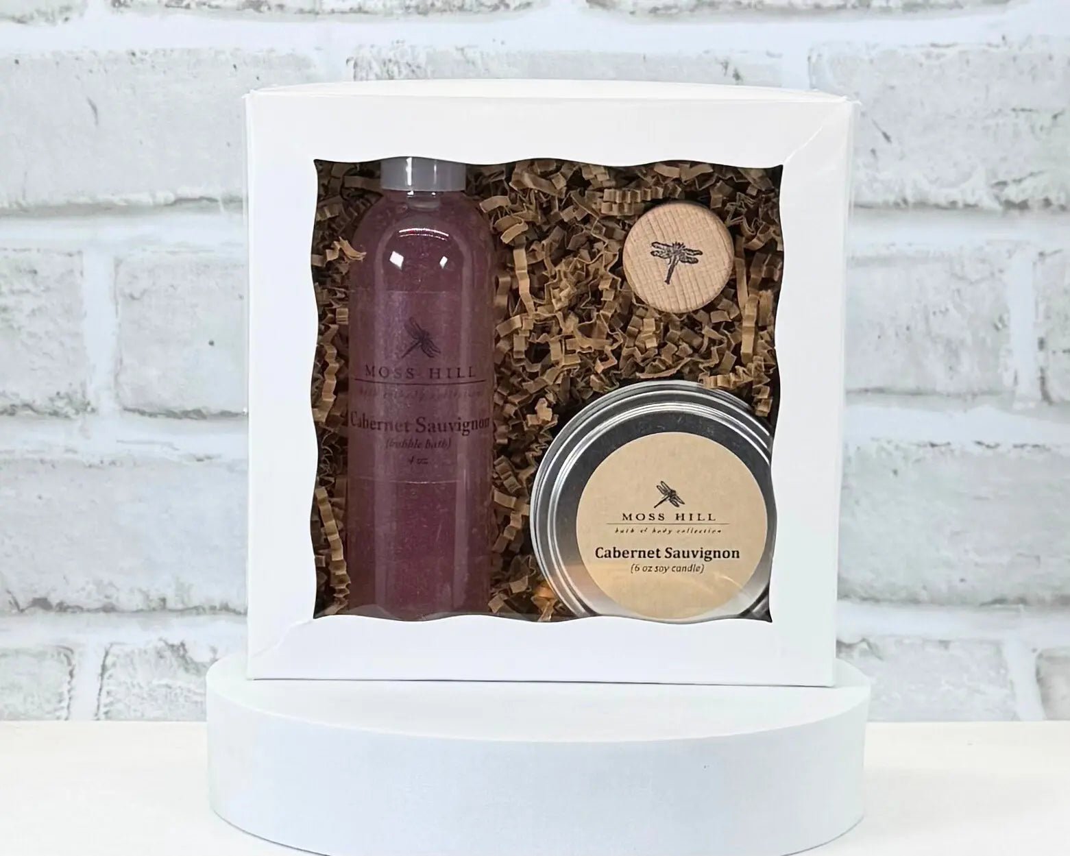 A Moss Hill body care gift set with handmade bubble bath and soy candle in the scent rose. Packaged in a white box on a white brick background.