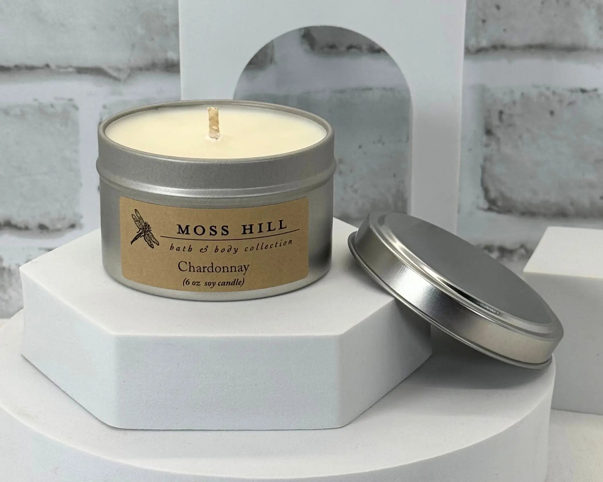 A soy candle in the scent Chardonnay. Hand-poured into a silver tin. Sitting on a white table top.