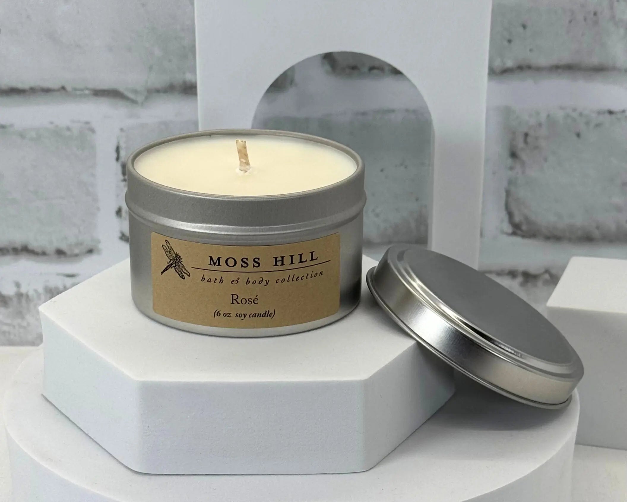 A soy candle in the scent rose. Hand-poured into a silver tin. Sitting on a white stand with a white background.
