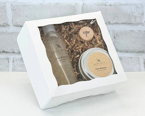 A Moss Hill body care gift set with a handmade bubble bath and soy candle in the scent Chardonnay. Wrapped in a white box placed on a surface with white brick background.