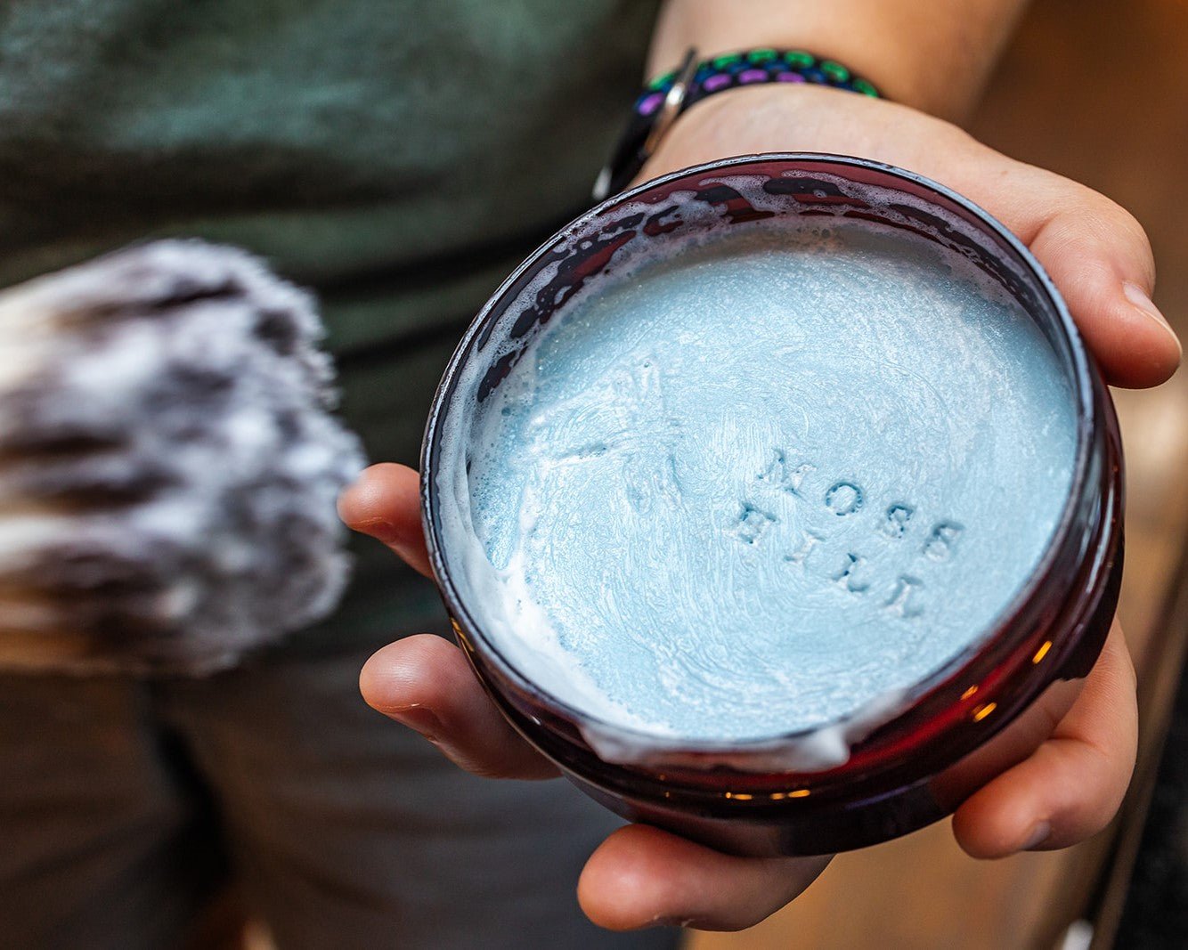 Brown container with blue shave soap with embossed details lathered up with a shave brush.