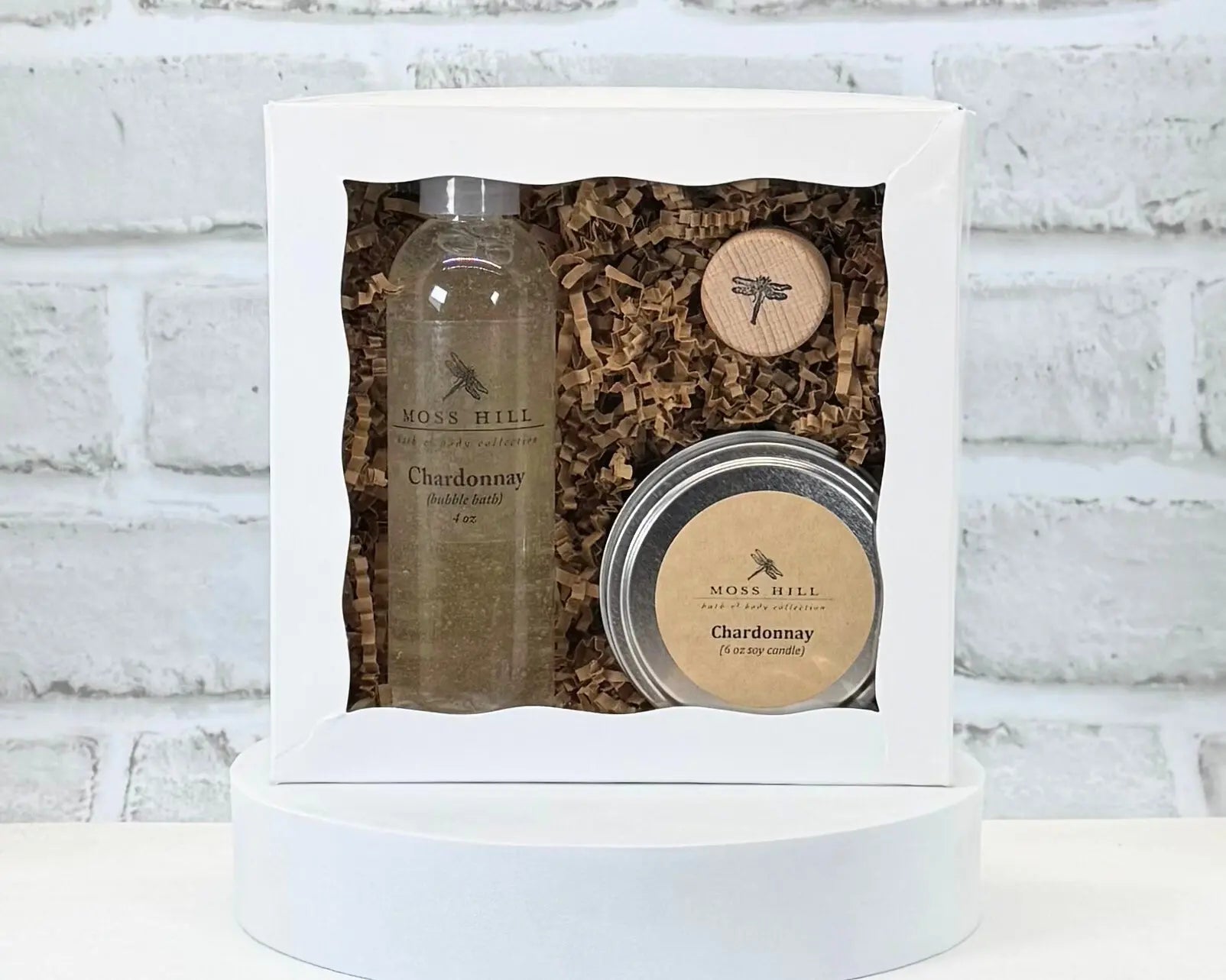 A Moss Hill body care gift set with handmade bubble bath,  soy candle, and wine cork in the scent Chardonnay. Packaged in a white box on a white brick background.