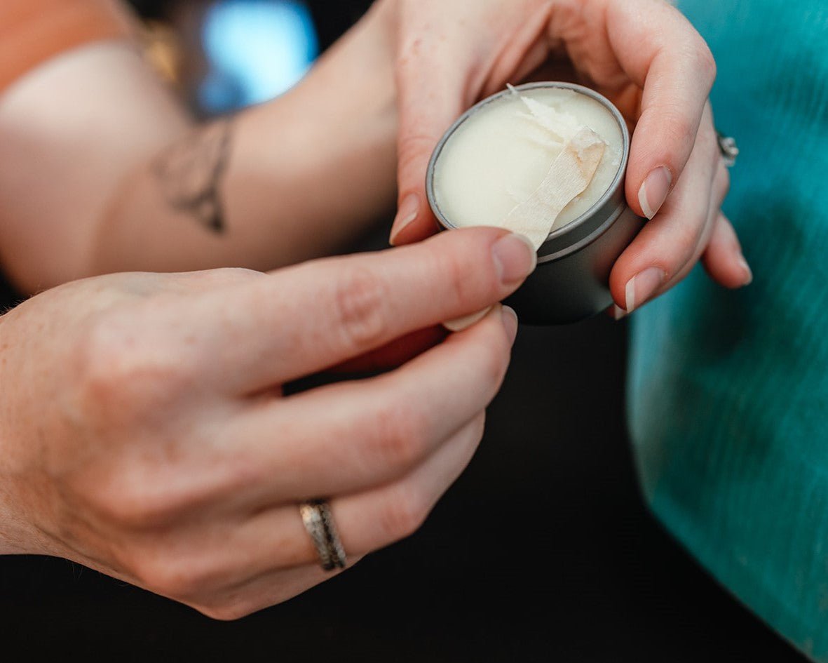 A hand holding a small wooden spoon to scoop out white dry skin relief cream to use on hands.