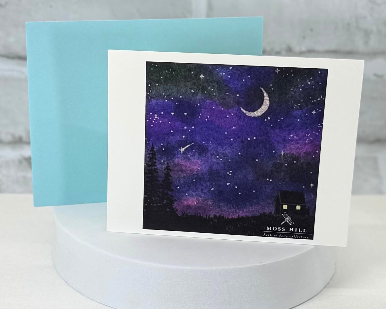 A watercolor greeting card with a starry night design and 'Moss Hill' branding on a white stand. Representing the Sweet Dreams sent profile for handmade bath and body products.