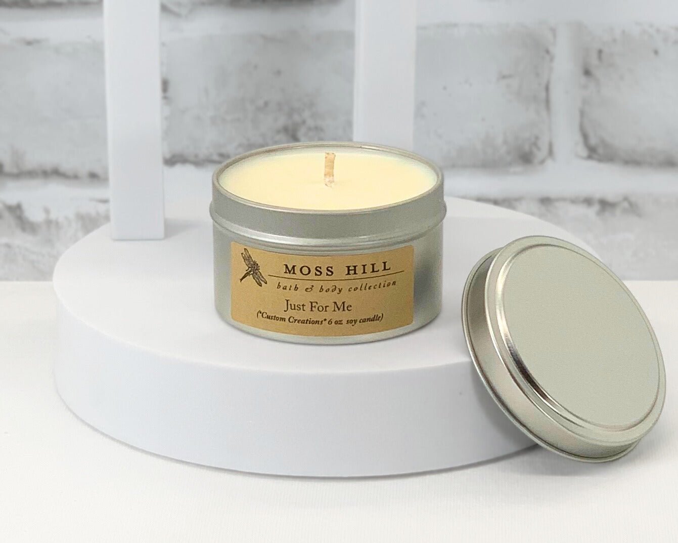A custom 6 oz candle by Moss Hill in a metallic tin with a label, lid off to the side, on a white surface with a brick background.