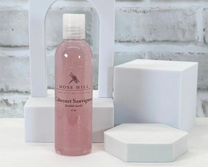A pink bottle of handmade bubble bath in the scent Cabernet Sauvignon. Place on a white surface with a white background.