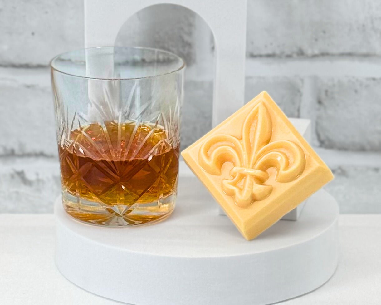 Bar soap shaped as the fleur de lis in the scent Bathe in Bourbon sitting next to a bourbon glass on a white table.