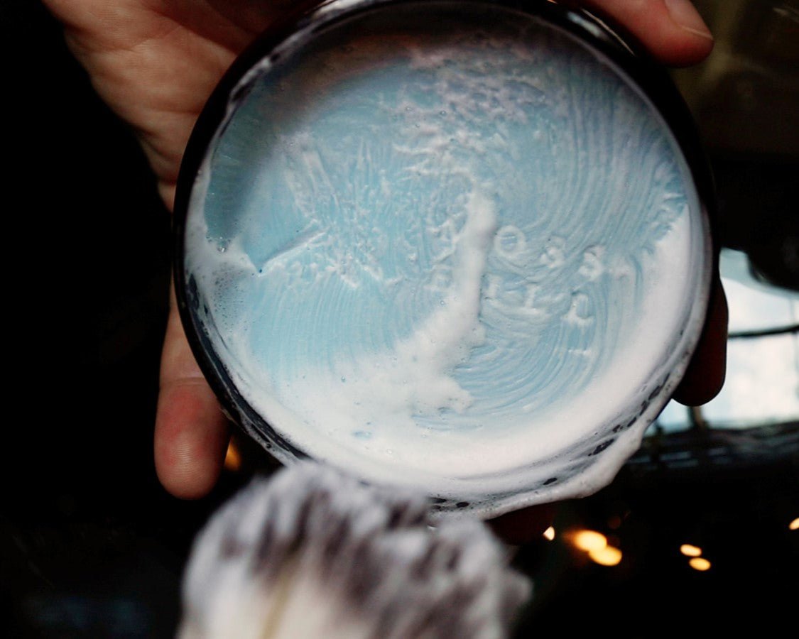 Handmade shave cream lathered up in a clear brown container being held in one hand.